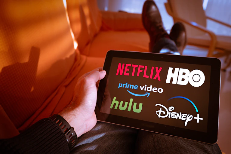 A man holding a tablet displaying the Netflix, Hulu, Amazon prime video, HBO and Disney+ logos on screen.