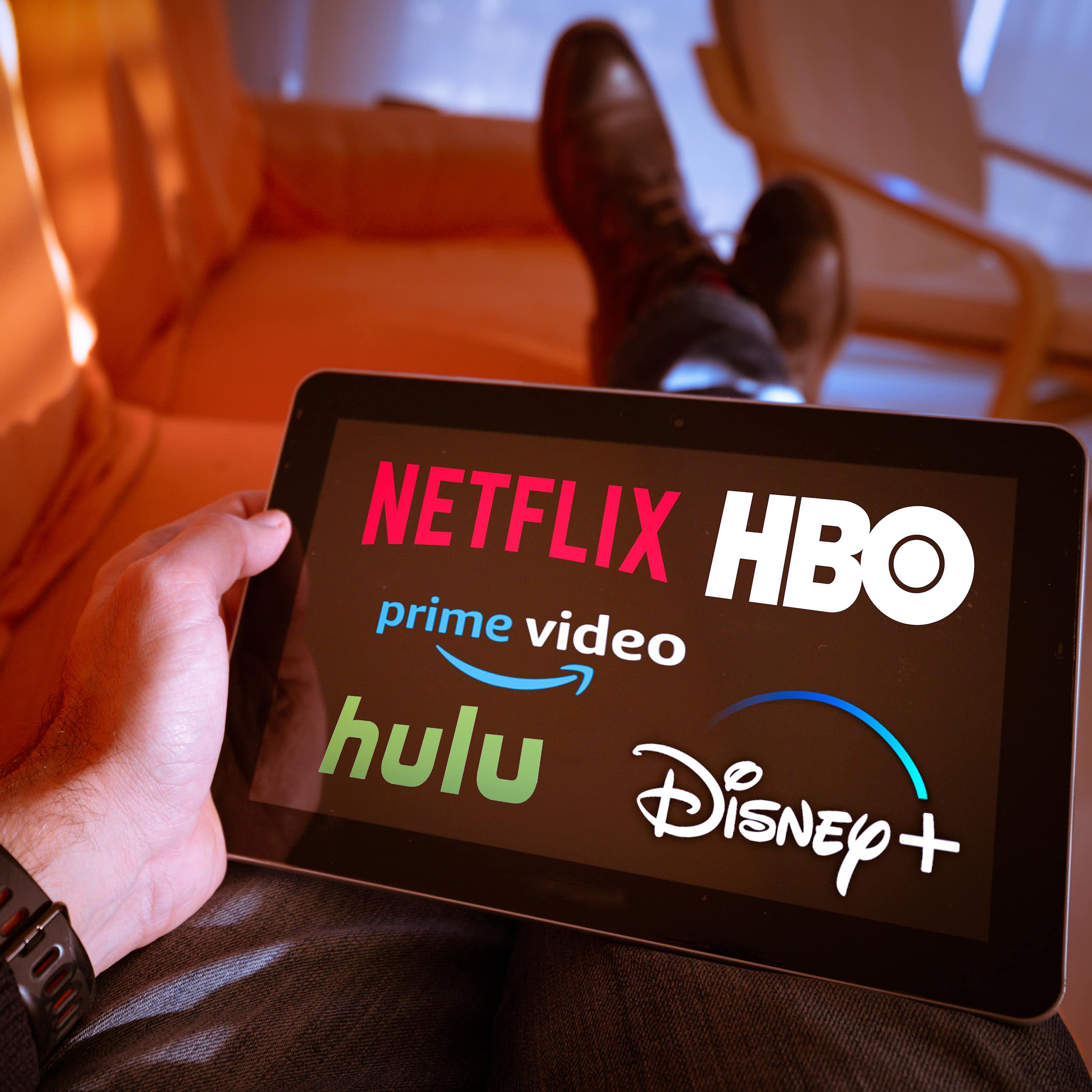 A man holding a tablet displaying the Netflix, Hulu, Amazon prime video, HBO and Disney+ logos on screen.