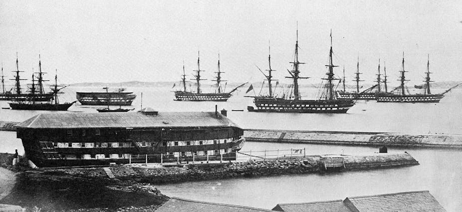 Archival black and white photo of eight ships in a harbour, a large prison ship is in the foreground