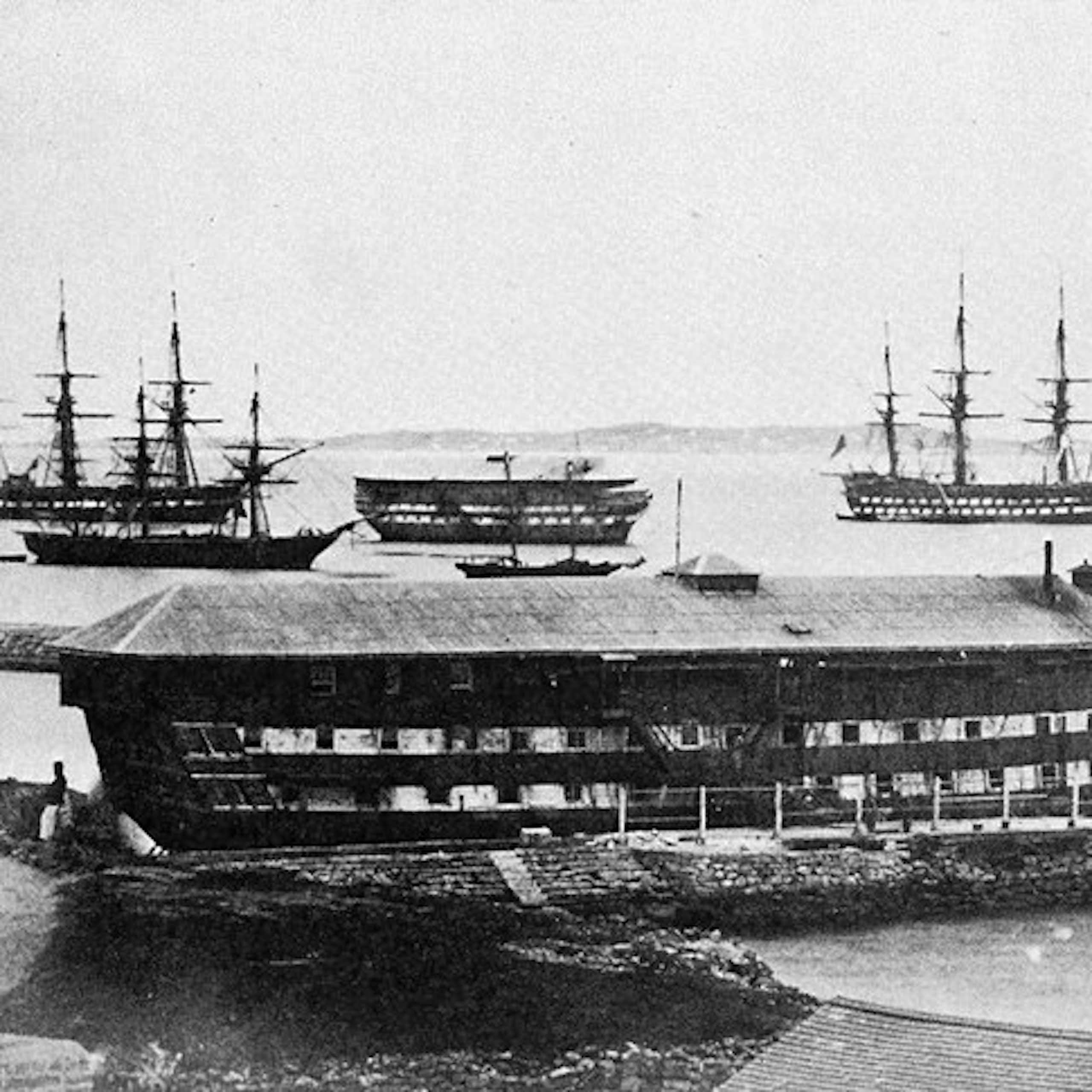 Archival black and white photo of eight ships in a harbour, a large prison ship is in the foreground