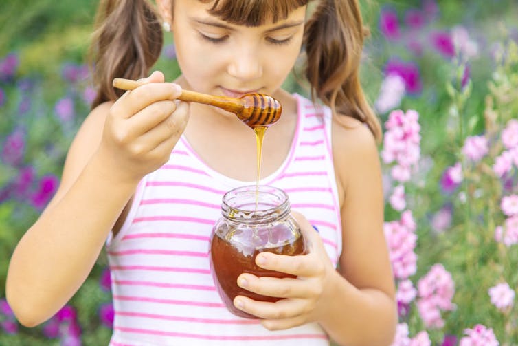 A young girl standing outside in a field of flowers holds a jar and a spoonful of honey.