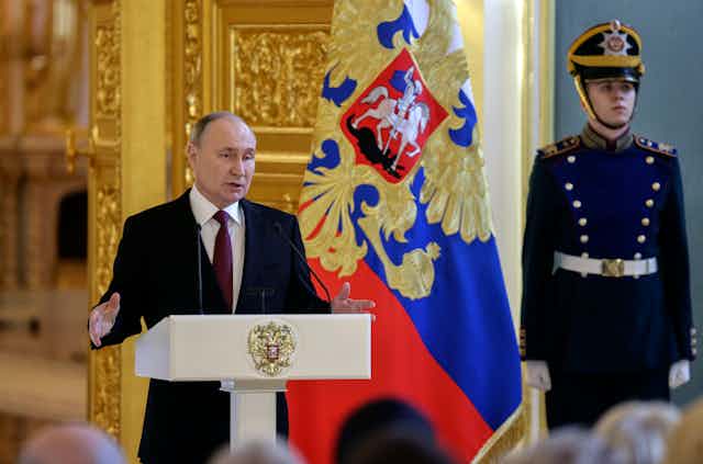 Russian president Vladimir PUtin delivers remarks from a lectern flanked by a soldier in ceremonial unifform.
