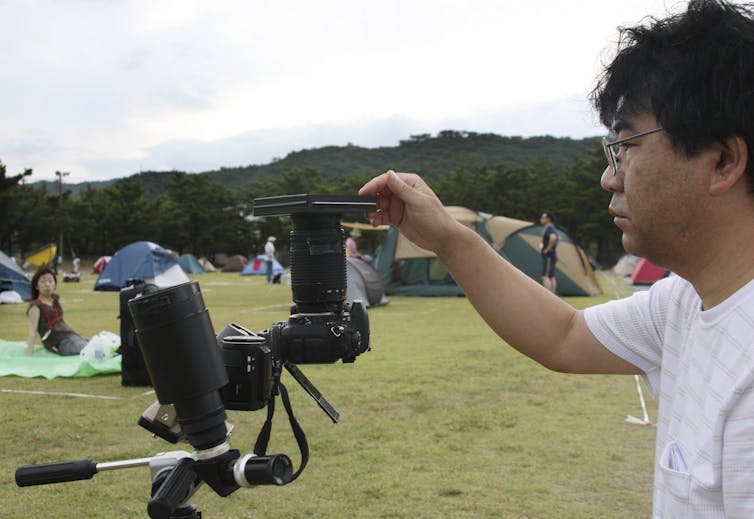 Tents under a grey sky and a person with a camera while another person lies on the ground.