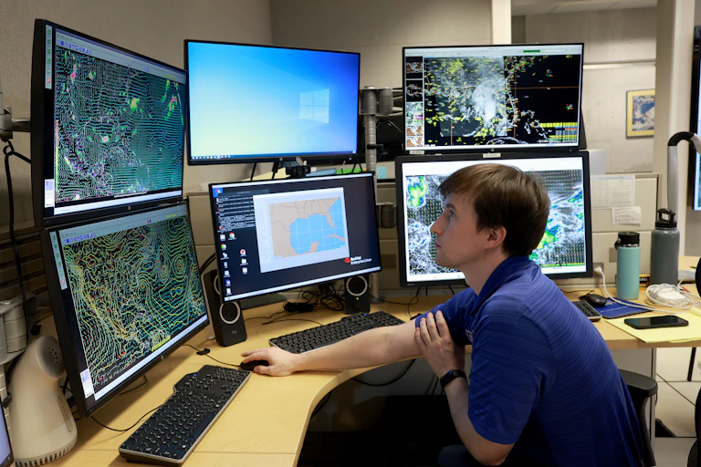 A person sits at a desk using a mouse and keyboard in front of several screens.
