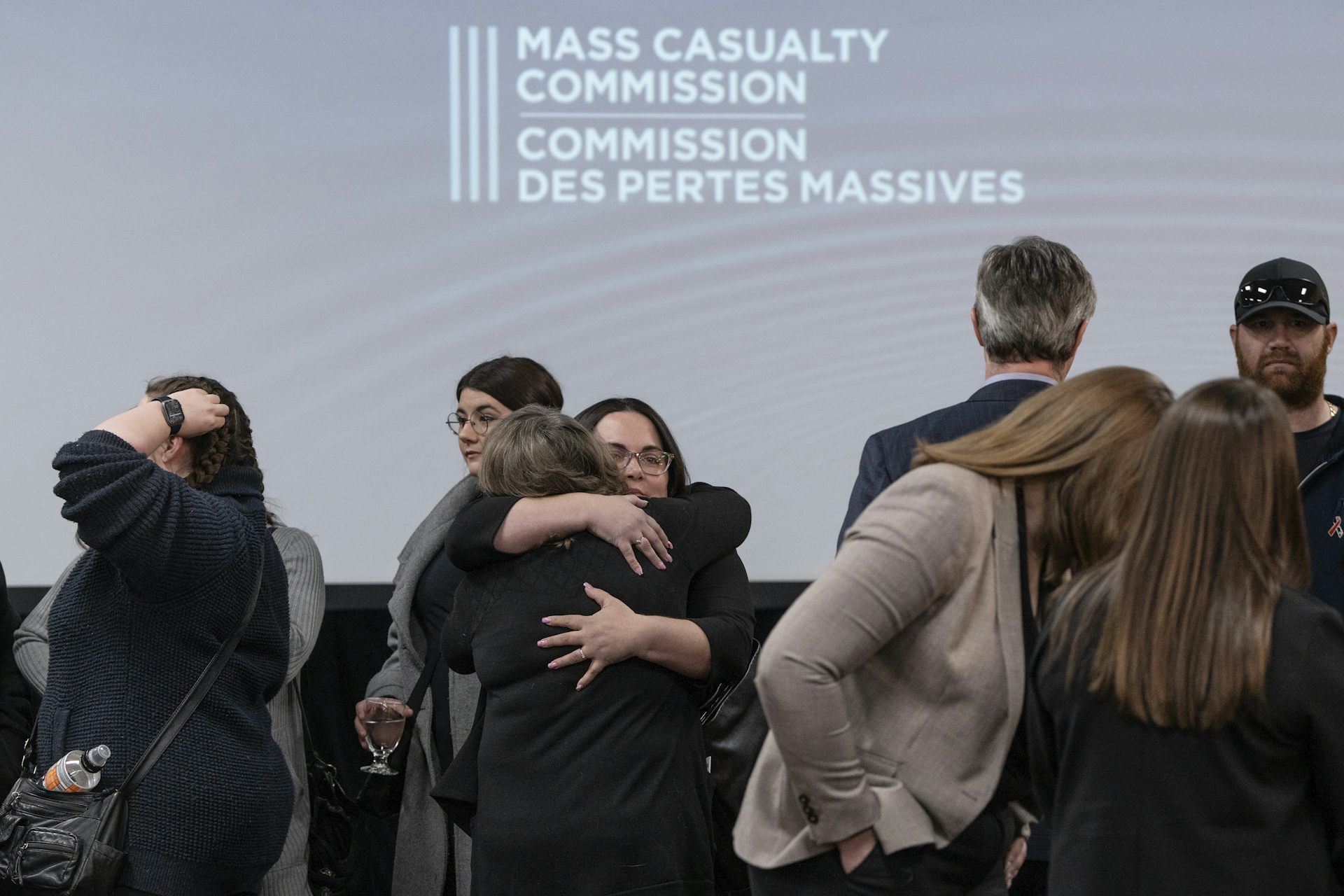 People embrace in front of a large screen that reads Mass Casualty Commission.