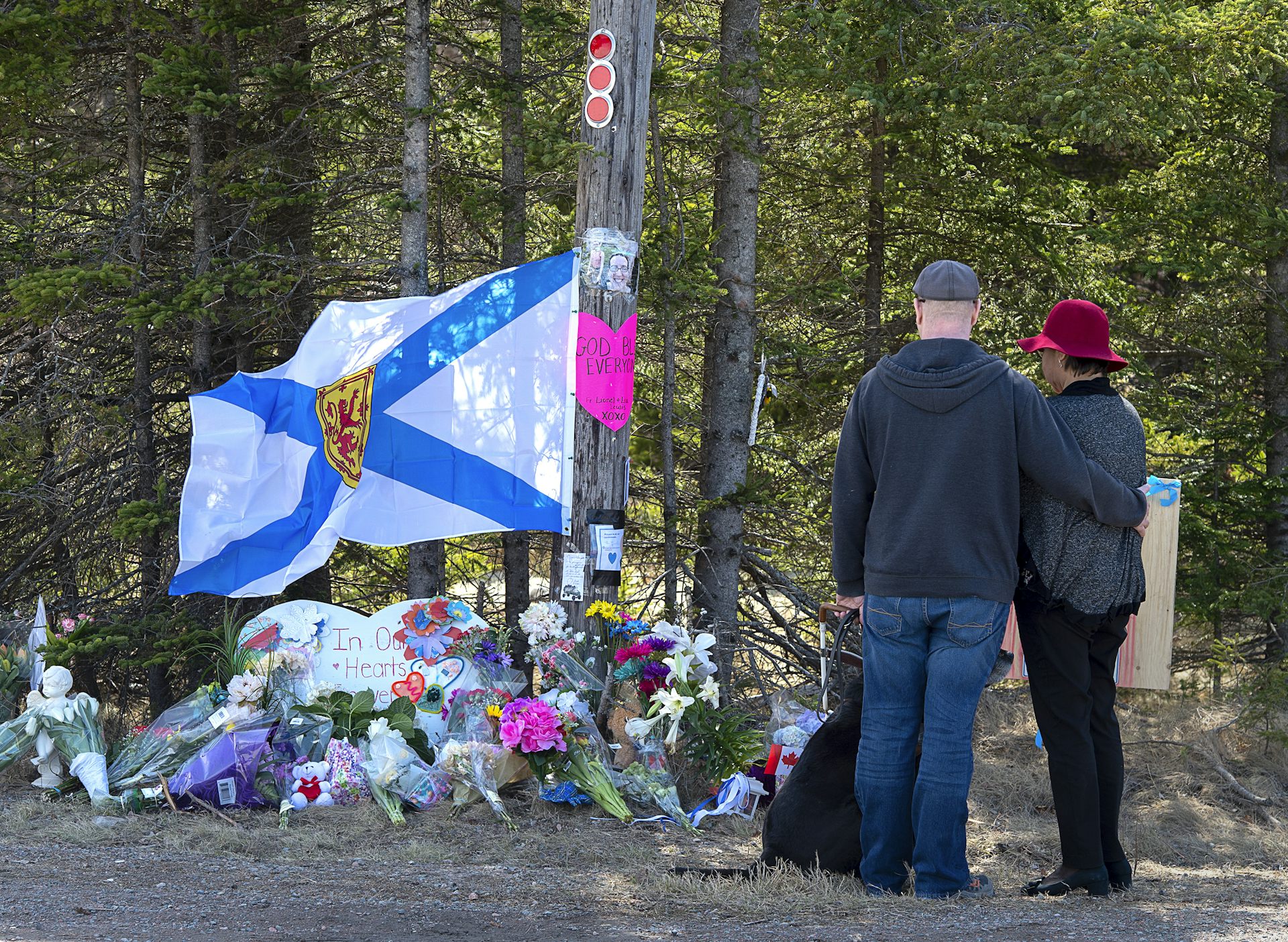 A couple and their dog look down at a makeshift shrine to shooting victims with a prominent blue and white Nova Scotia flag.