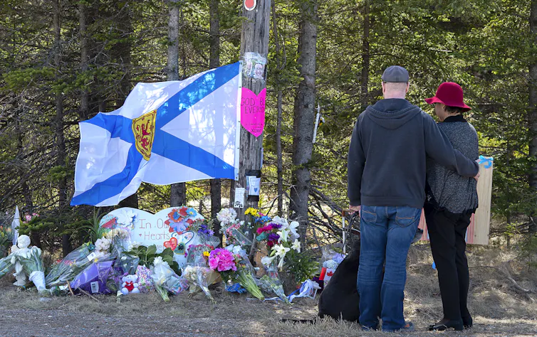 A couple and their dog look down at a makeshift shrine to shooting victims with a prominent blue and white Nova Scotia flag.