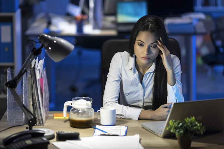 woman with bored expression holds left fingers to her left temple while staring down at her desk, which has a computer, coffee and a lamp on it