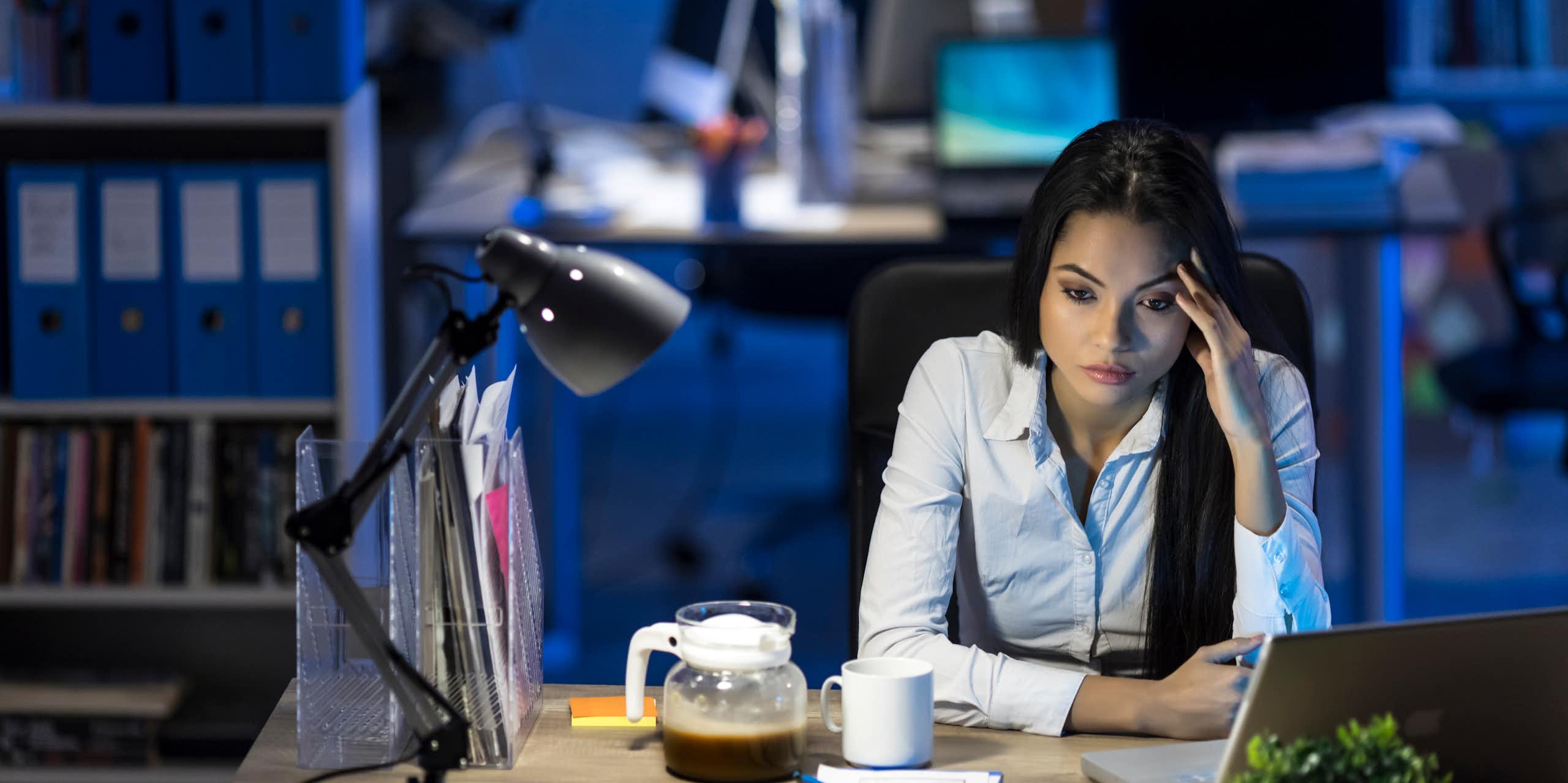 woman with bored expression holds left fingers to her left temple while staring down at her desk, which has a computer, coffee and a lamp on it