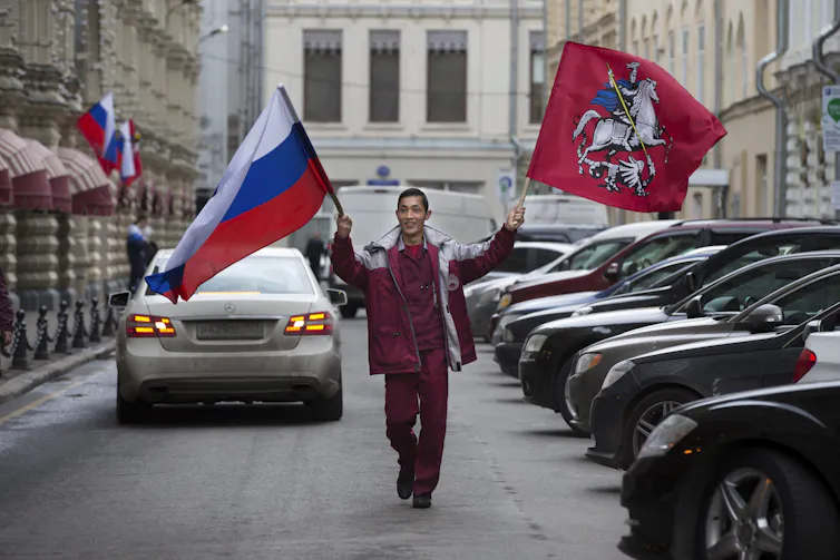 A smiling man carries the Russian and Tajikistan flags as he walks past a row of parked cars.