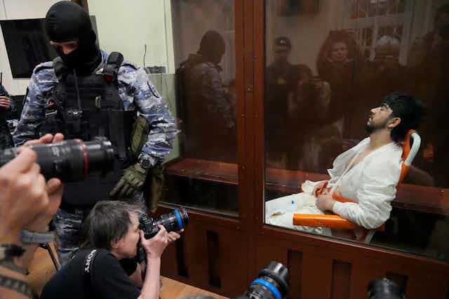A man in white prison garb sits in a glass cage as photographers take pictures of him.