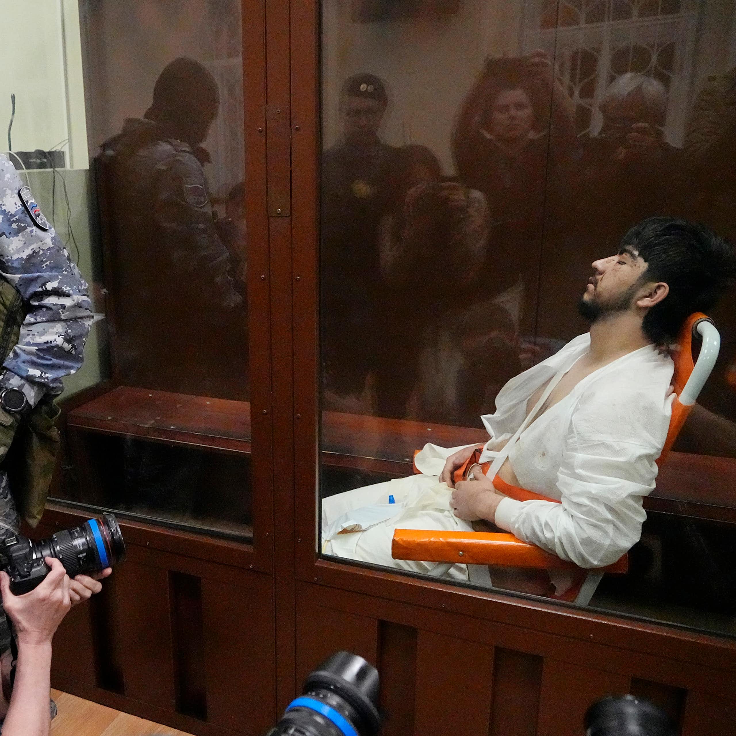A man in white prison garb sits in a glass cage as photographers take pictures of him.