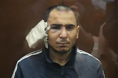 A man with a black swollen eye and a large bandage on his ear sits in a glass cage.
