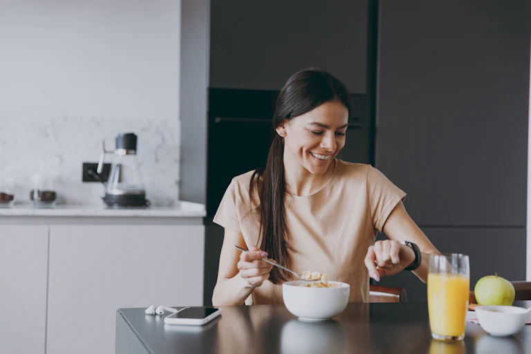 Uma mulher está na cozinha tomando café da manhã. Ela está olhando a hora no relógio de pulso.