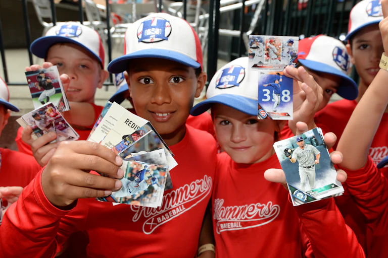A group of young fans wearing ballcaps red baseball uniforms hold up their baseball cards.