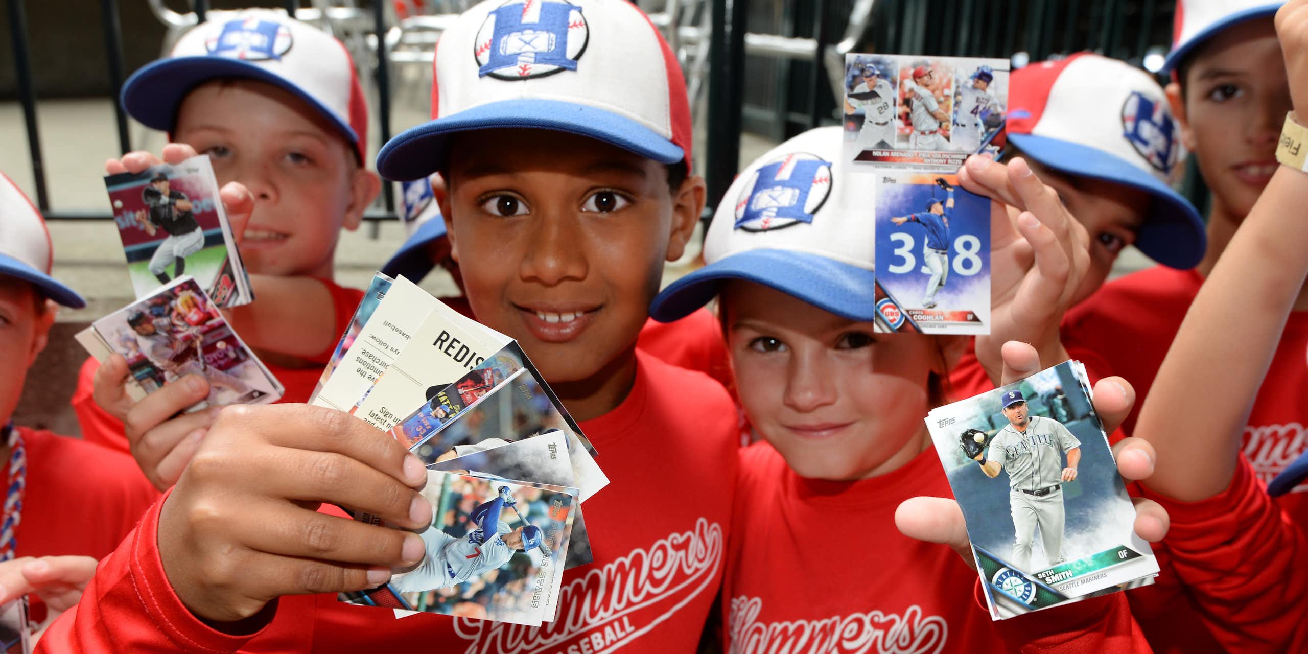 A group of young fans wearing ballcaps red baseball uniforms hold up their baseball cards.