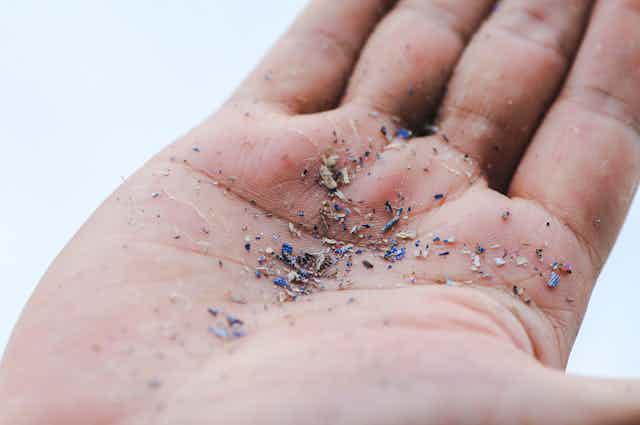 Uma imagem em close da mão de uma pessoa. Em sua palma, ela segura fragmentos de microplásticos