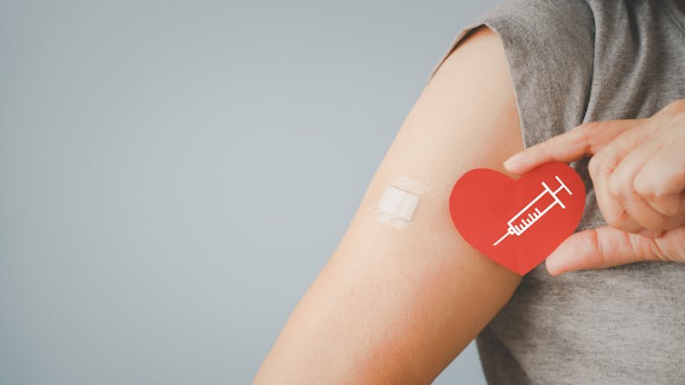 A person's arm and shoulder, with a small bandage on the upper arm and the other hand holding a heart with a syringe drawn on it