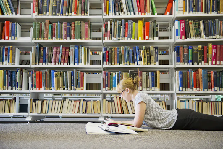 A young woman lies on the floor reading books in front of library book shelves.