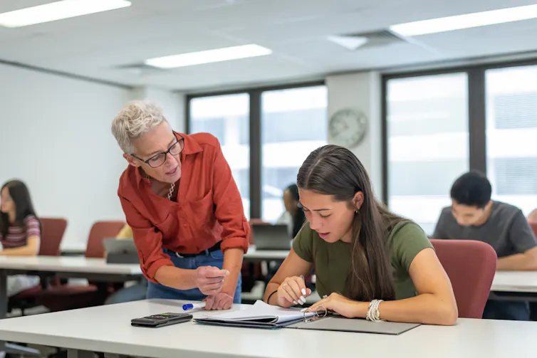 A woman leans over the desk of a student working, talking to the student.