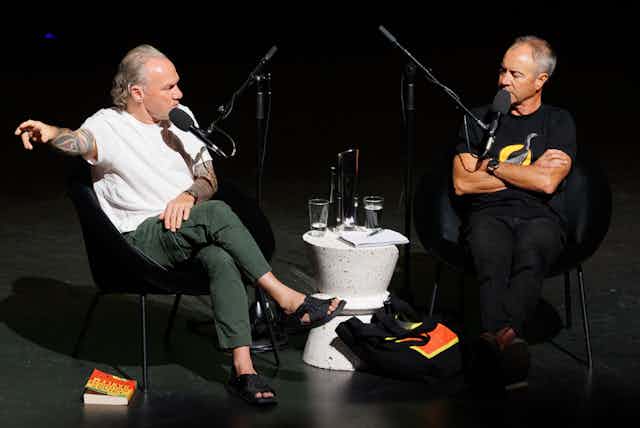 Two men sittiing on a black stage talking.