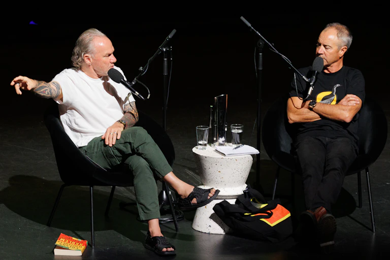 Two men sittiing on a black stage talking.