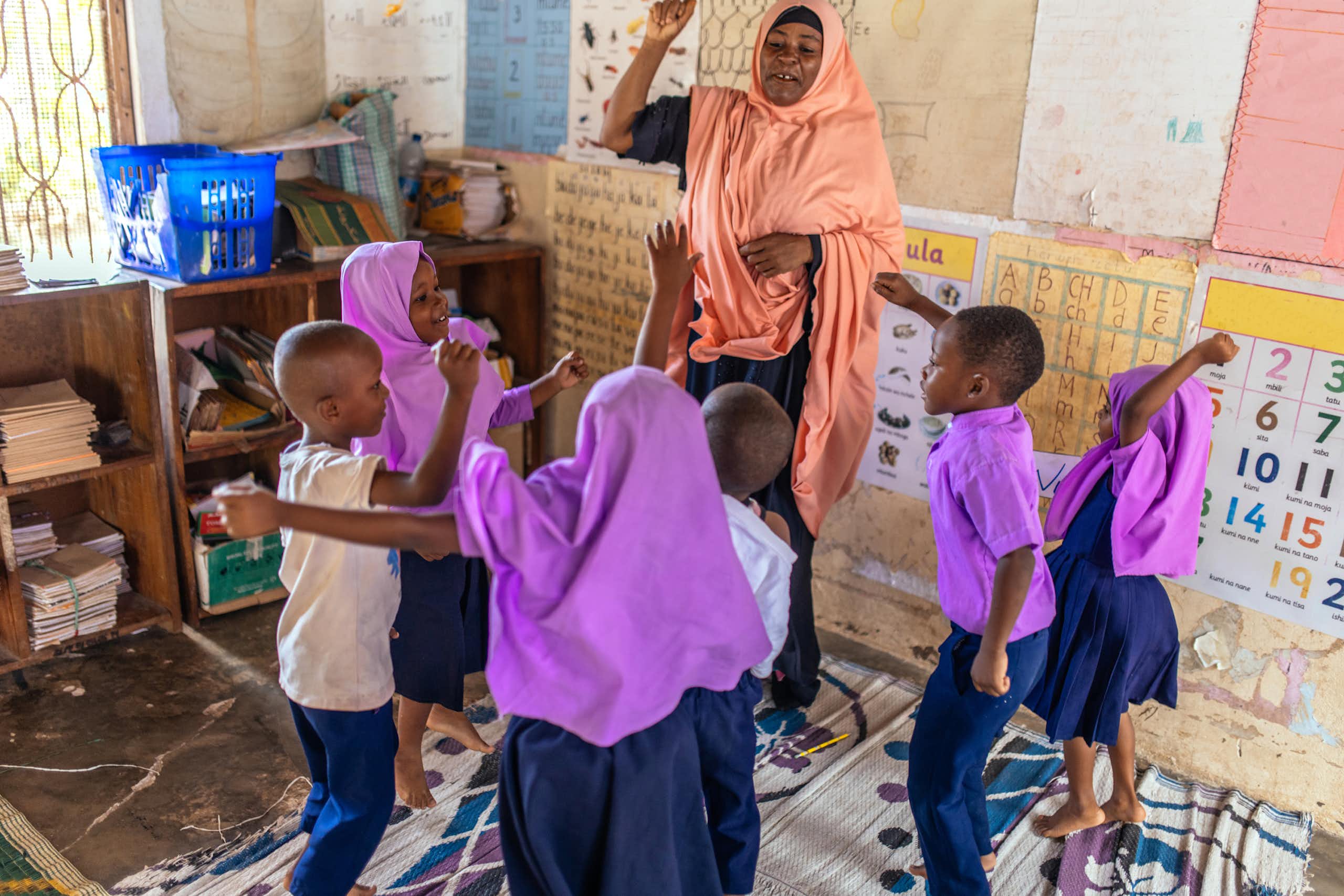 An adult woman in a peach-coloured hijab and black dress dances with a small group of schoolchildren in a classroom