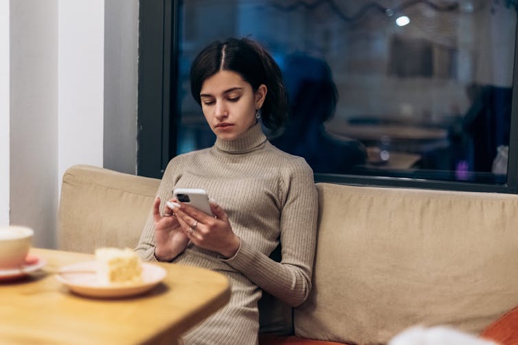 A young woman sitting alone at a cafe with a slice of cake, scrolling on her phone.