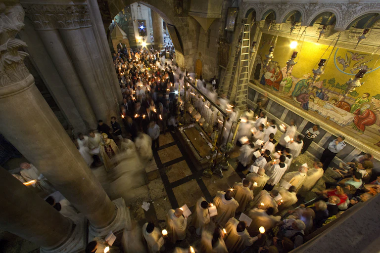 Men in white priestly robes walk in a procession while holding candles through a hallway with ornate pillars and a wall with paintings depicting followers surrounding Jesus' body.