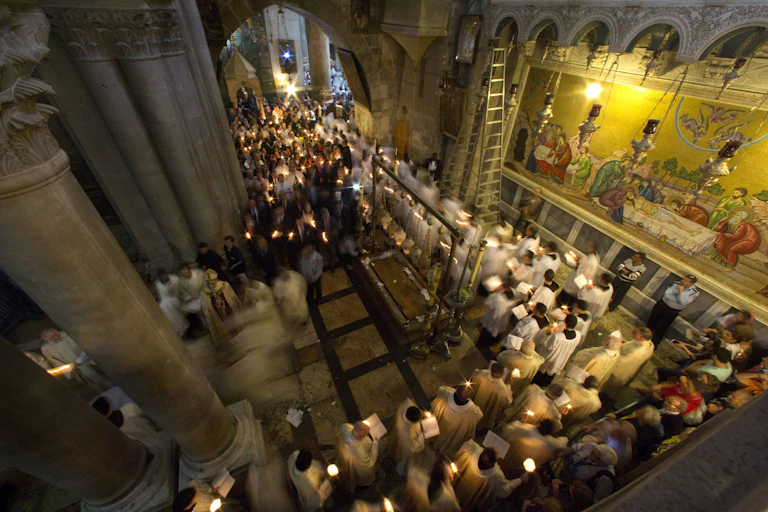 Men in white priestly robes walk in a procession while holding candles through a hallway with ornate pillars and a wall with paintings depicting followers surrounding Jesus' body.