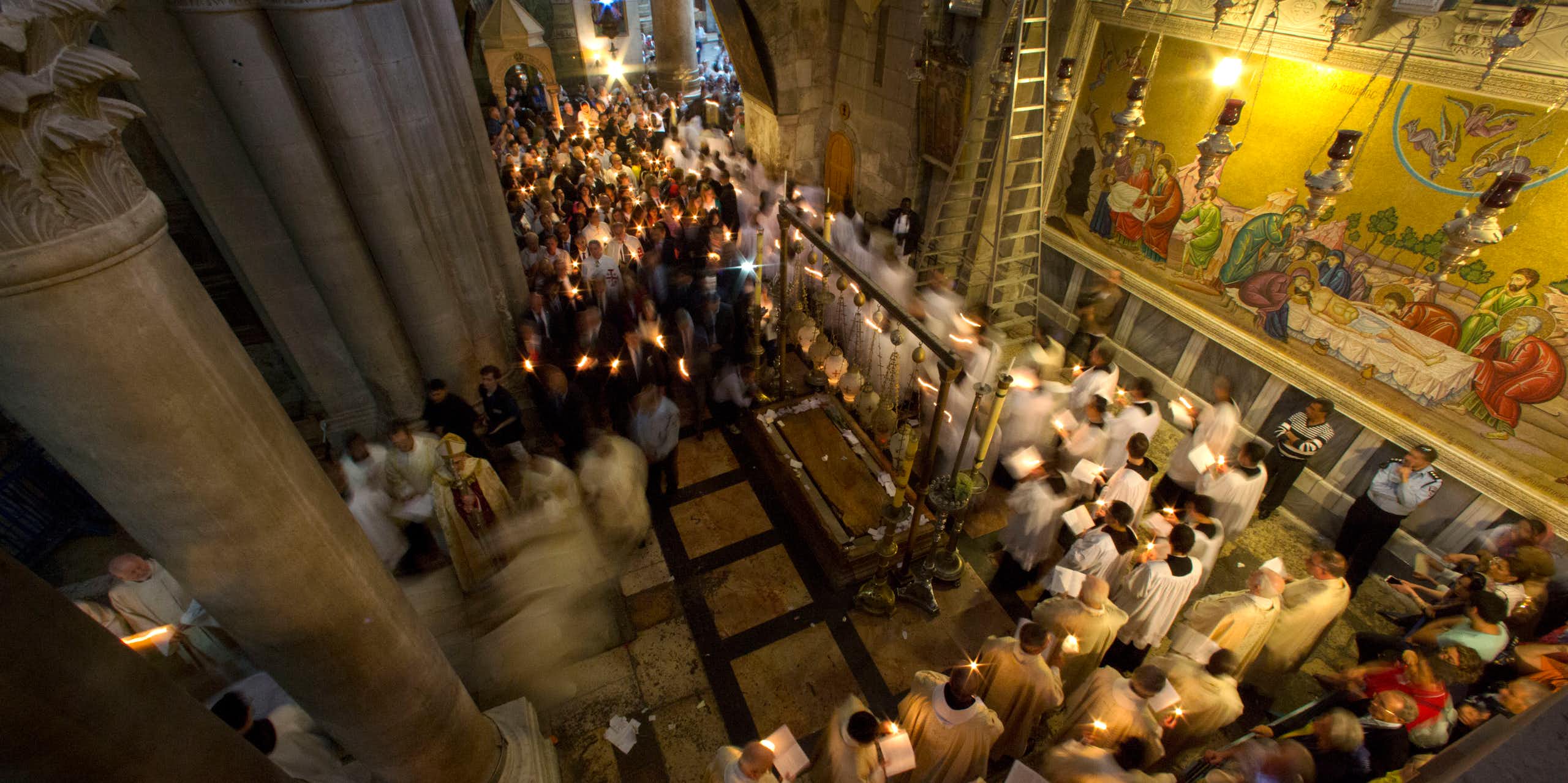 Men in white priestly robes walk in a procession while holding candles through a hallway with ornate pillars and a wall with paintings depicting followers surrounding Jesus' body.