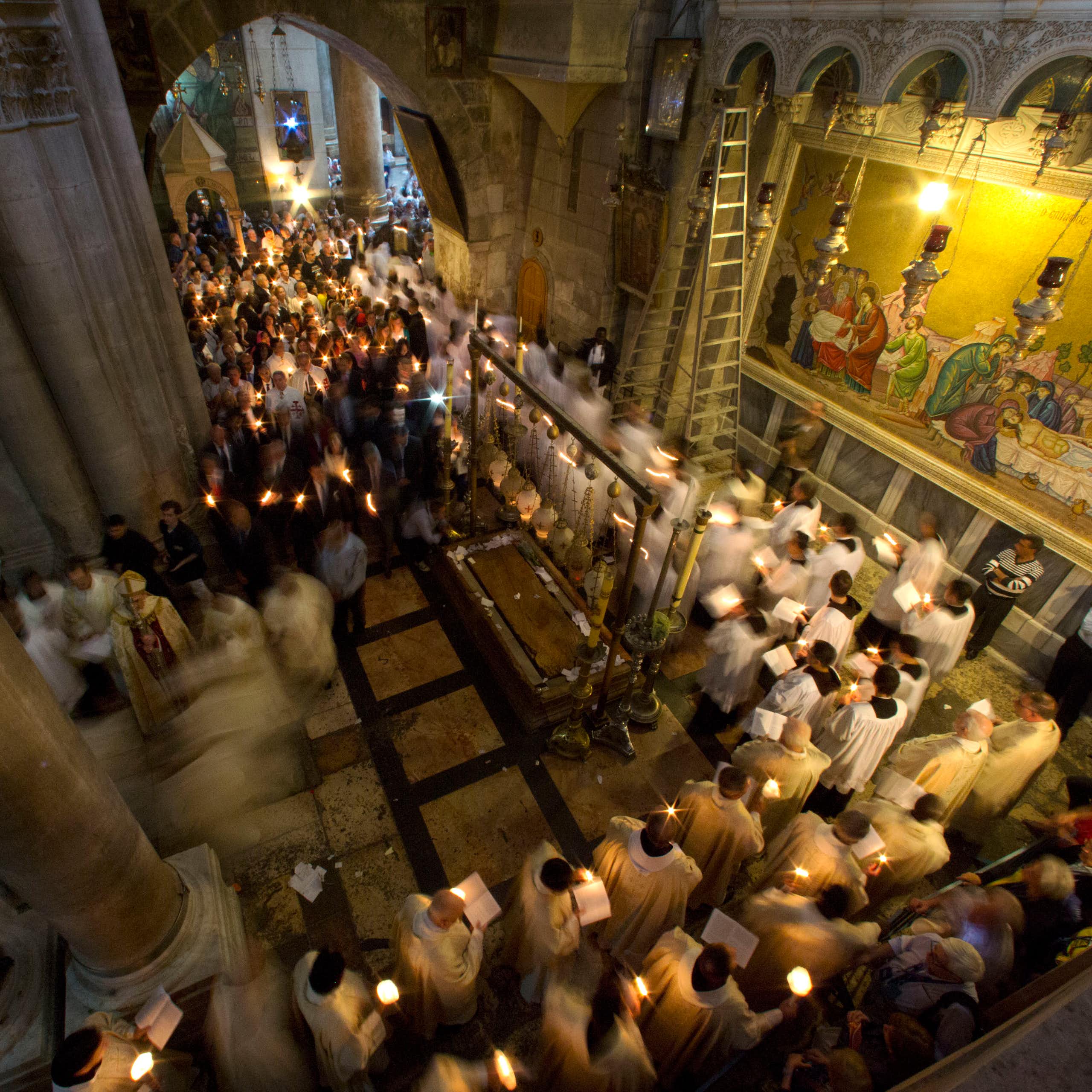 Men in white priestly robes walk in a procession while holding candles through a hallway with ornate pillars and a wall with paintings depicting followers surrounding Jesus' body.