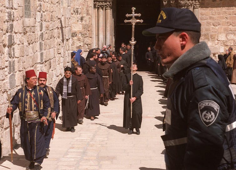 Men wearing long green garbs walk in a procession and one in the center holds a tall crucifix.