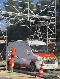 Un pompier recouvert d’une combinaison nettoie un camion en l’aspergeant d’une substance blanche.
