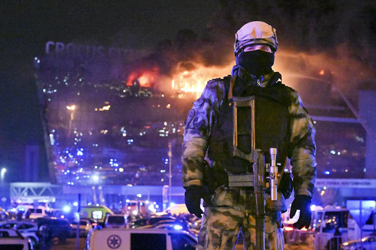 Un soldat se trouve au premier plan de la photo tandis qu'un bâtiment en flammes se trouve à l'arrière-plan.