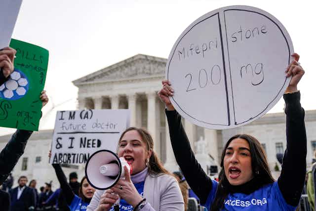 Women hold a bullhorn and a sign that says 'Mifepristone' outside the Supreme Court on a gray day.