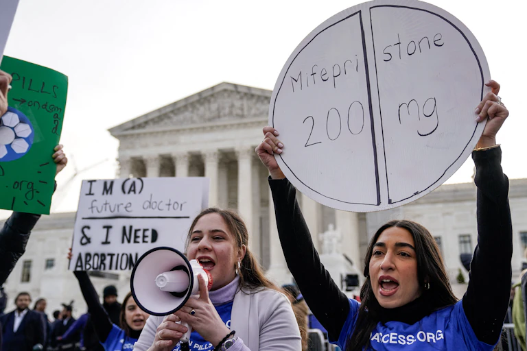Women hold a bullhorn and a sign that says 'Mifepristone' outside the Supreme Court on a gray day.