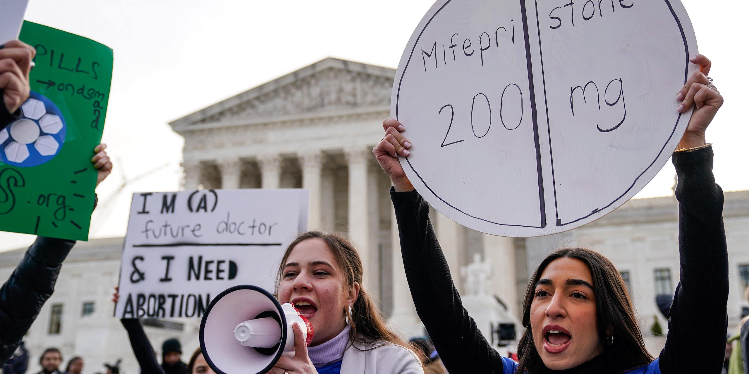 Women hold a bullhorn and a sign that says 'Mifepristone' outside the Supreme Court on a gray day.