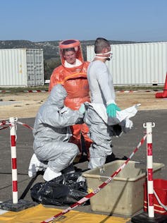 Trois pompiers sont en combinaison de protection. L’un des trois est en train d’aider un autre à enlever sa combinaison.