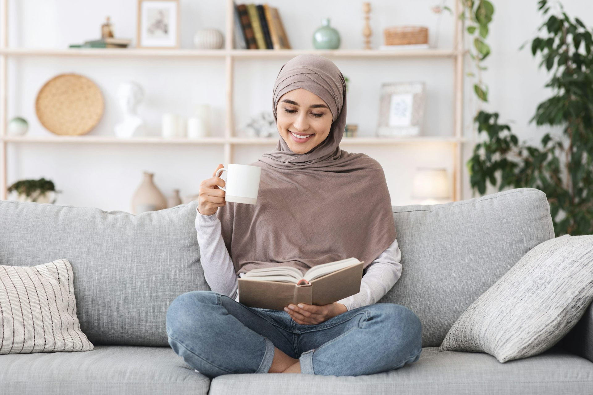 A woman sitting on a sofa holding a mug in one hand and a book in the other