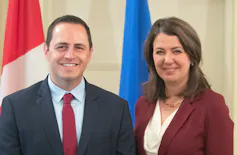 Two people seen smiling in business clothes in front of a red and white flag and a blue flag.