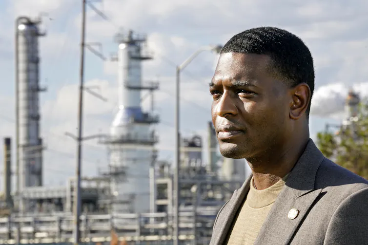 A man in a sport coat stands in front of a refinery.