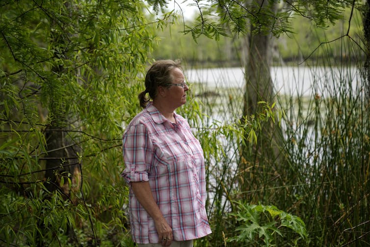 A woman stands beside a large lake surrounded by plants, trees and tall grasses.