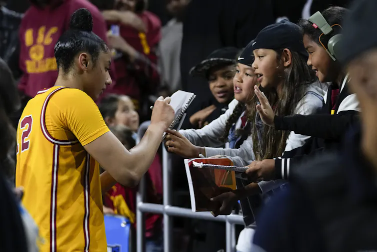 A young woman in a basketball uniform signs autographs for a group of young girls