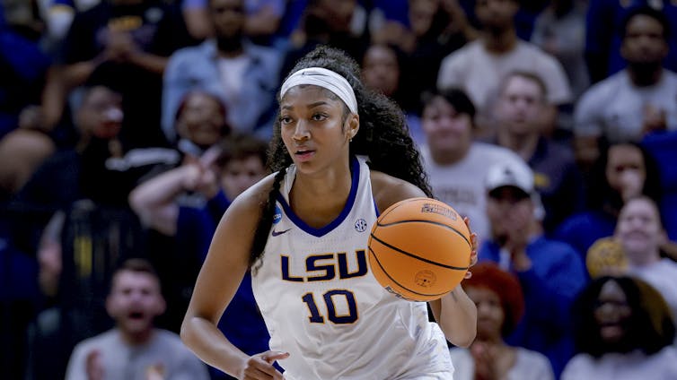 A young Black woman dribbles a basketball down a court while a crown watches on