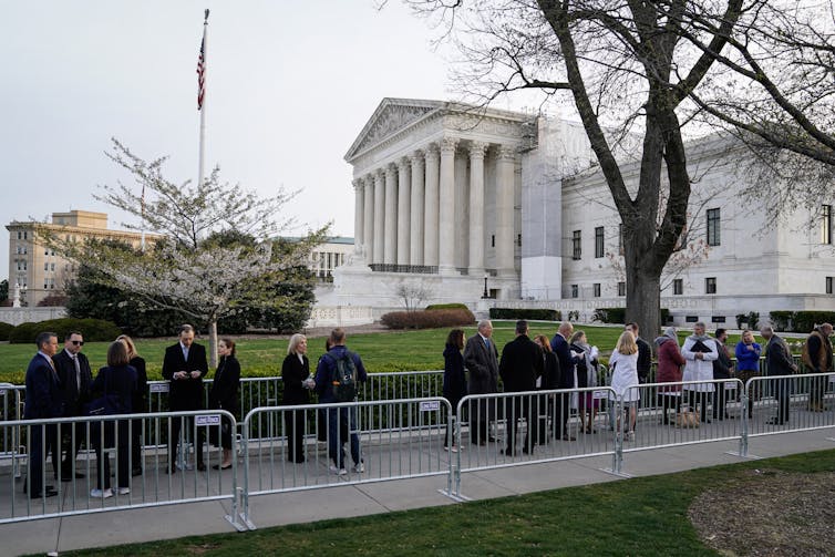 A line of people in formal clothing are seen behind barricades outside the Supreme Court on a grey day.
