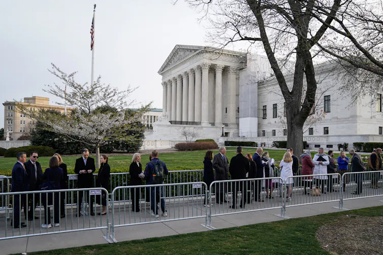 A line of people in formal clothing are seen behind barricades outside the Supreme Court on a grey day.