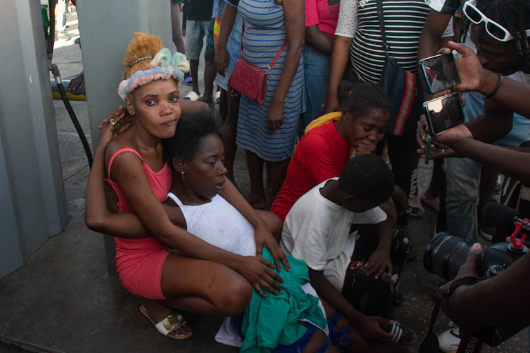 Au premier plan, une femme accroupie soutient une autre femme allongée. Derrière, on aperçoit une foule de gens.