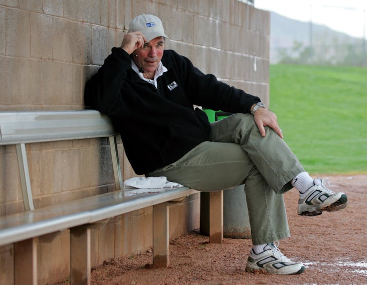 A middle-aged man sits cross-legged on a metal bench, tugging at the brim of his baseball cap.