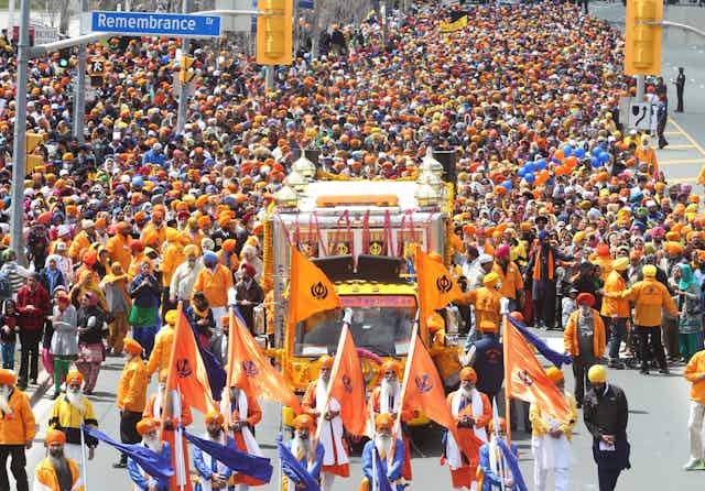 Hundreds walking in a parade, behind a bus with a temple-like structure atop and several  men in front of it, holding saffron-color flags.